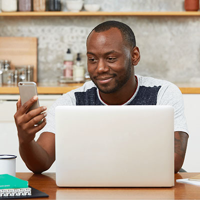 a man sitting at a table with a laptop computer smiling at a smart phone